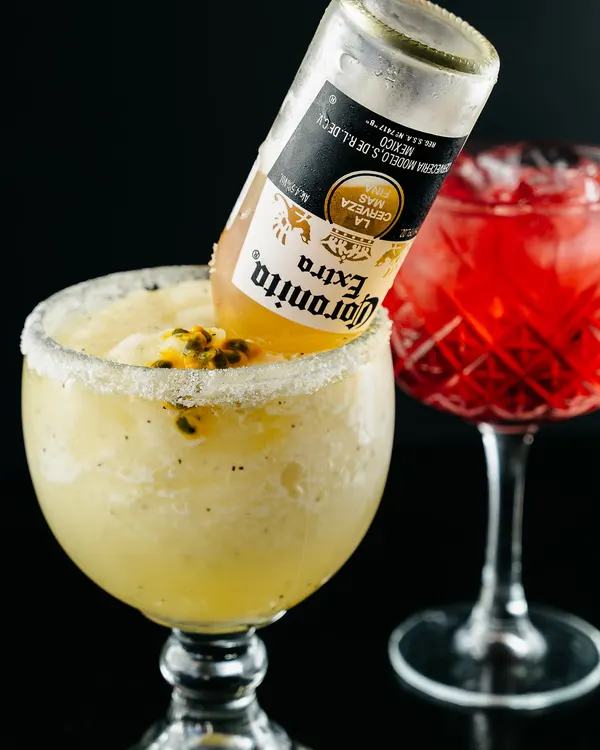 Margarita frozen con borde de sal y una botella de Corona invertida dentro, junto a un cóctel rojo en copa labrada.