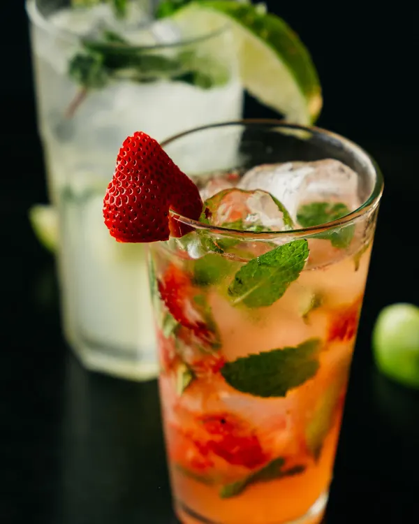Vaso alto con bebida rosada, hierbabuena y una fresa en el borde; detrás, una limonada con limón.