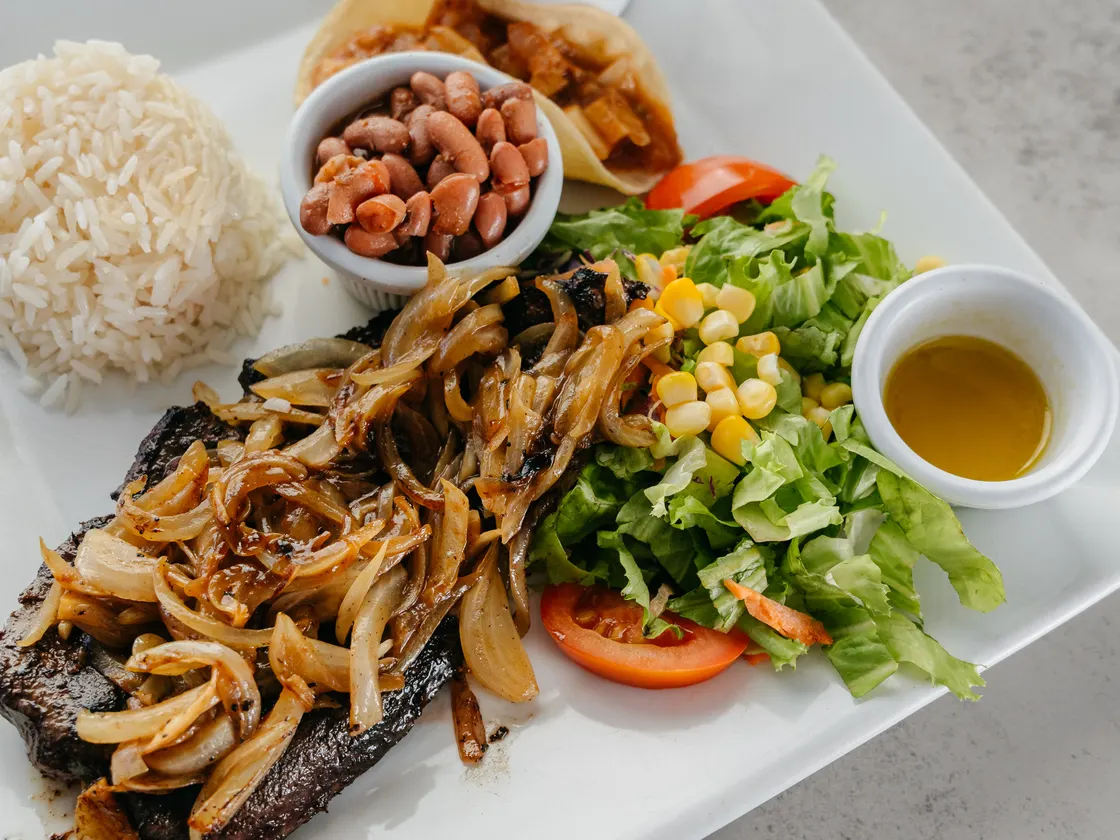 Carne a la plancha cubierta de cebolla salteada, con arroz blanco, frijoles, ensalada con maíz y tomate, y una salsa aparte.