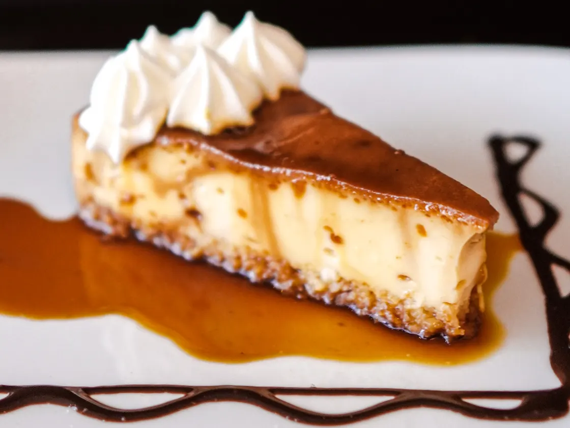 Tajada de cheesecake con crema batida encima, sobre caramelo y un hilo de chocolate.