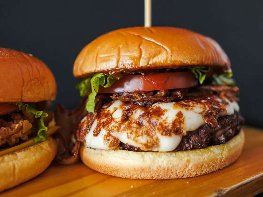 Hamburguesa alta con carne, queso derretido, tomate y lechuga en pan dorado, sobre una tabla de madera.