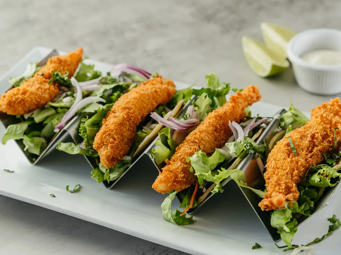 Cuatro tacos con tiras empanizadas, lechuga y cebolla morada en una base metálica, con limón y una salsa blanca al lado.